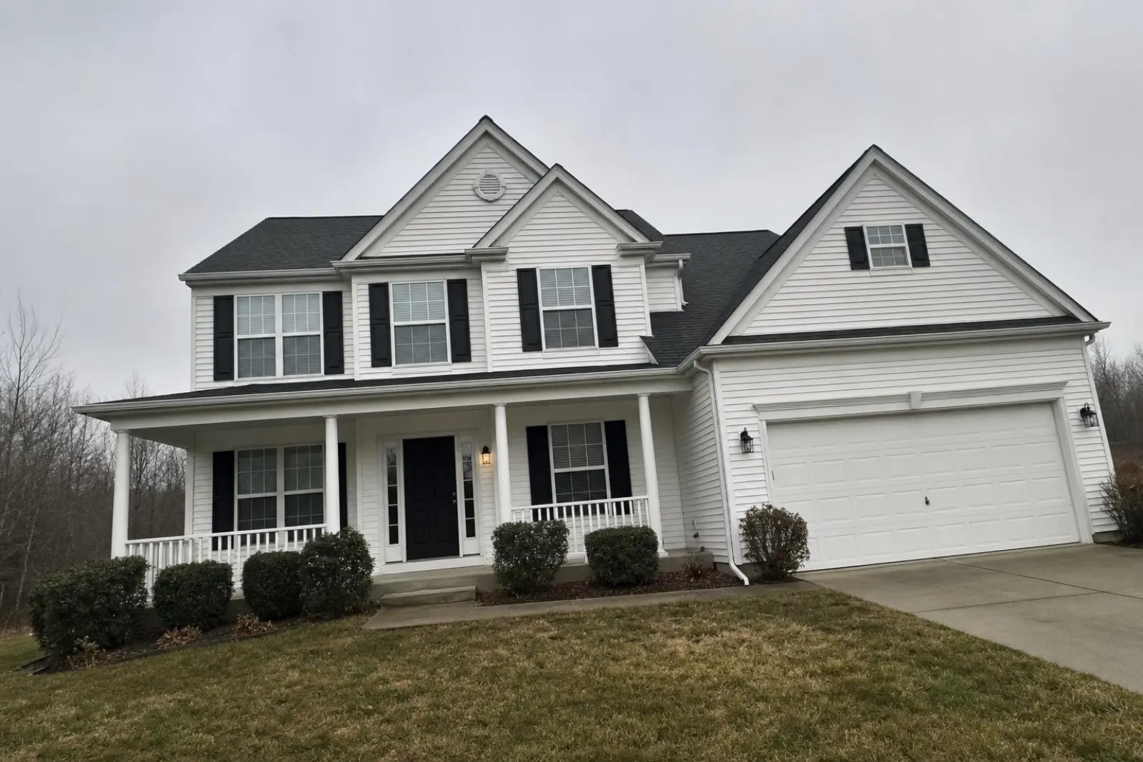 Two-story white house with black shutters, a front porch and a two-car garage under a flat grey overcast sky, shot at a slight tilt