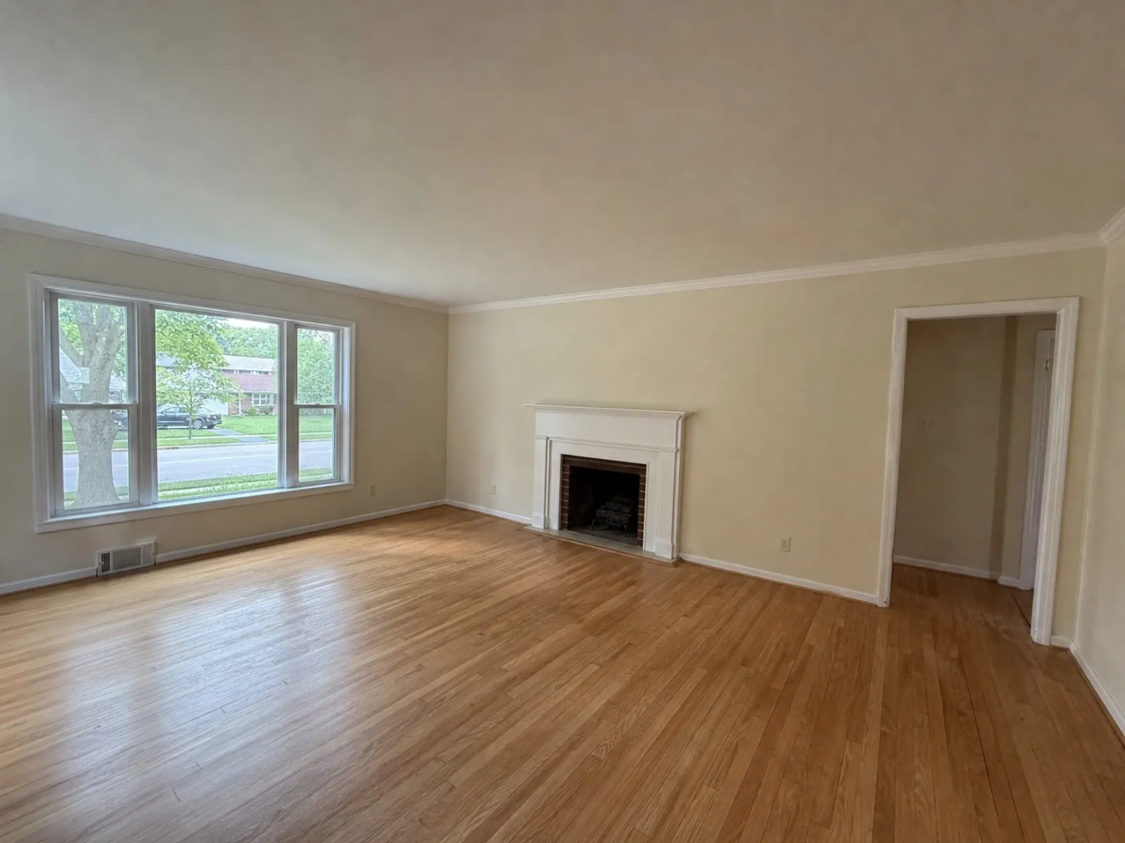Empty living room with oak floors, a white-mantel brick fireplace, a triple window facing the street and an open doorway on the right