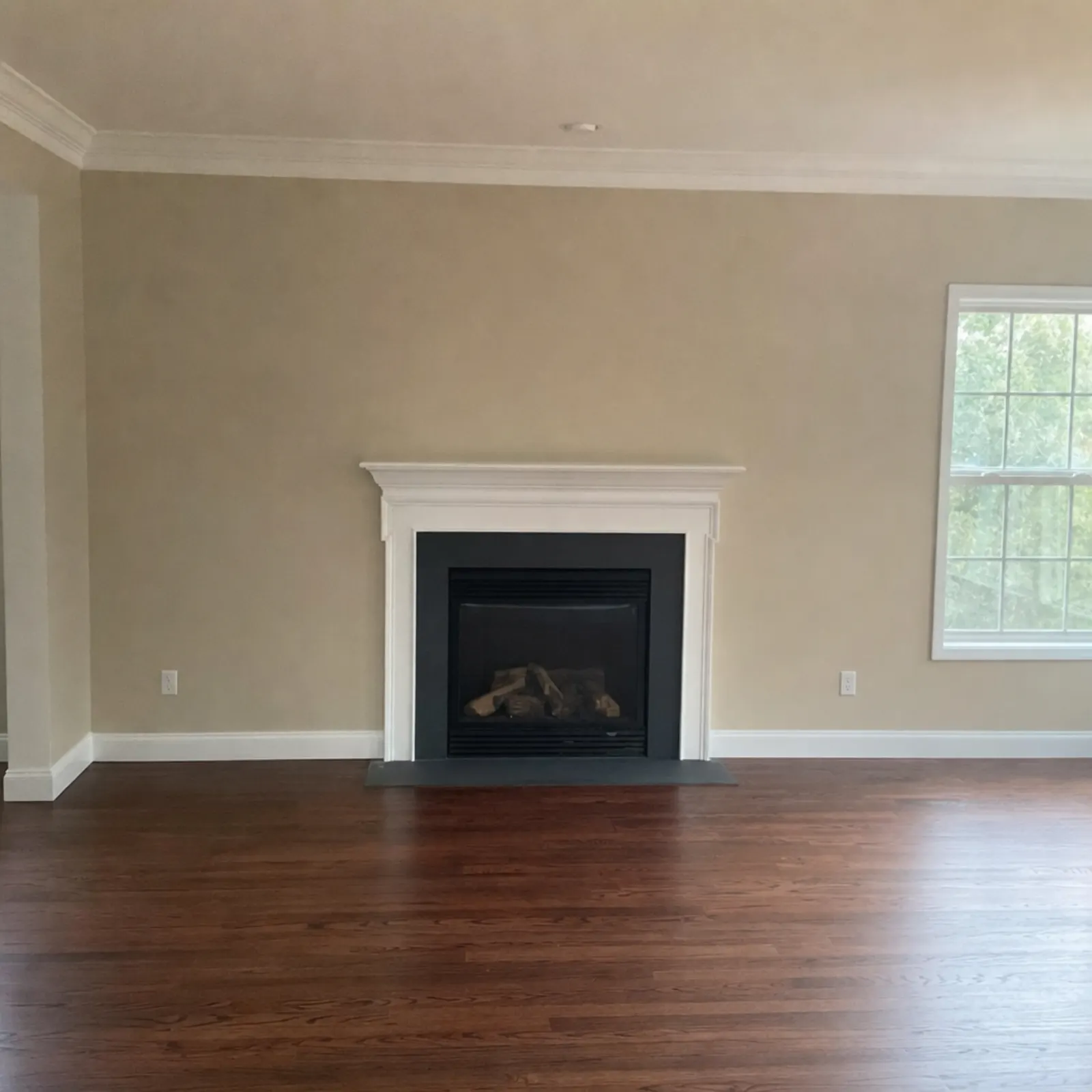 Empty living room with tan walls, dark hardwood floors, a white mantel with a black fireplace surround and a window on the right