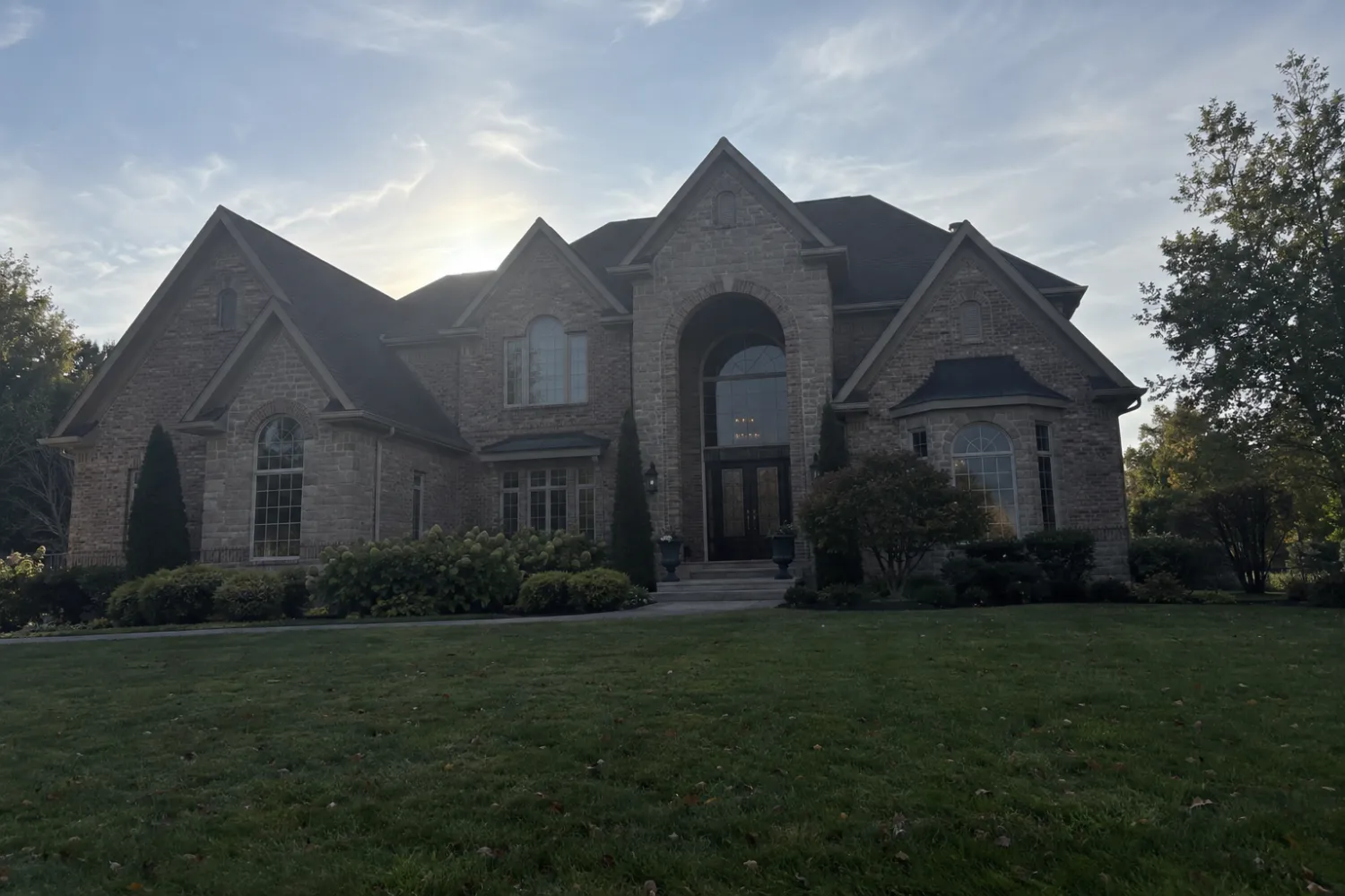 Large brick and stone house shot from the front lawn with the sun behind the roof, leaving the facade dark and flat